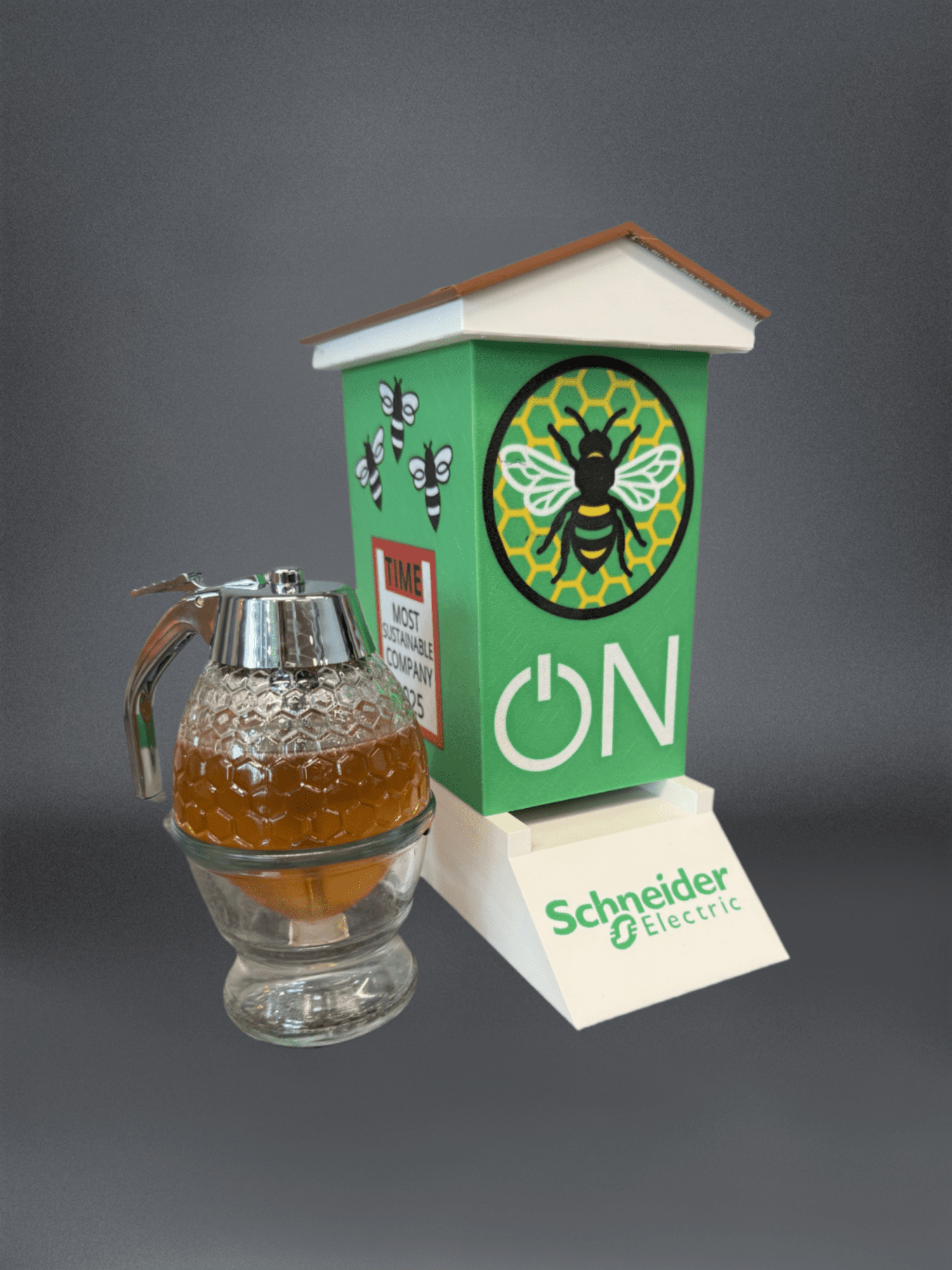 Desktop-scale Schneider Electric branded beehive model beside a glass honey dispenser