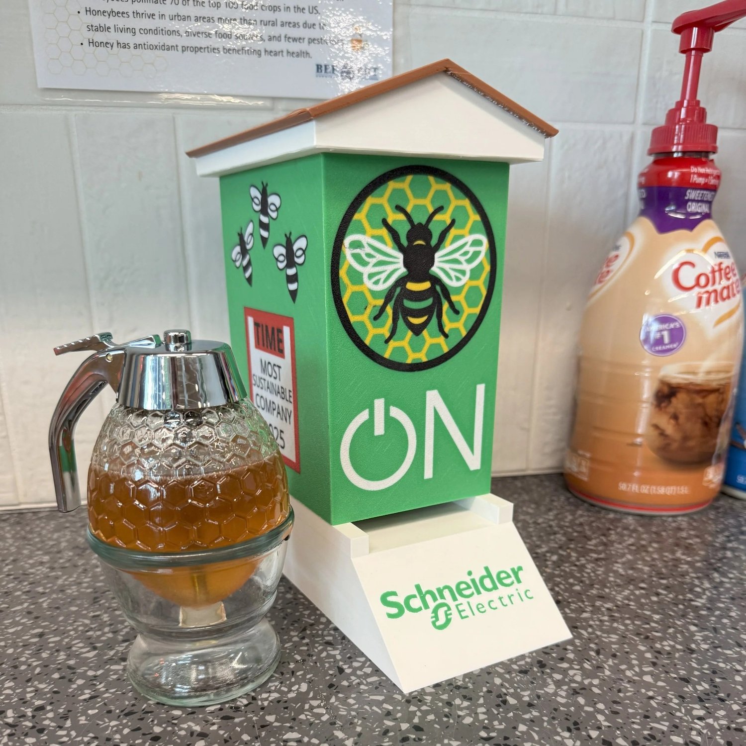 3D printed Schneider Electric beehive model on a counter beside a honey dispenser